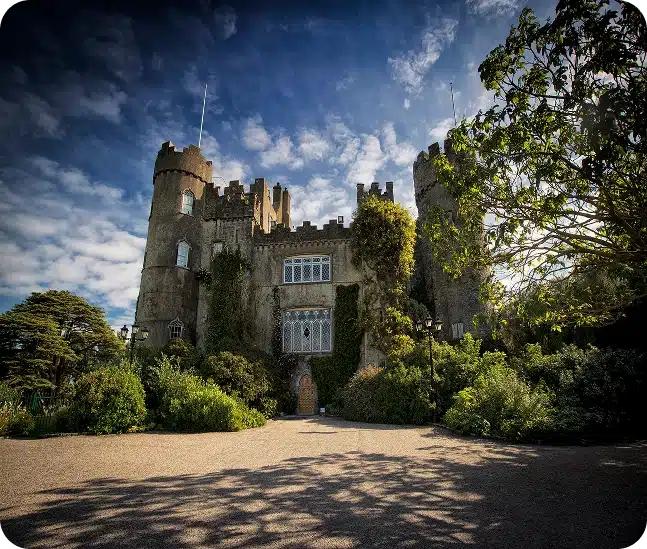 A large stone castle with tall towers and battlements sits surrounded by lush greenery and trees under a partly cloudy blue sky. Sunlight casts shadows on the ground in front of the castle.