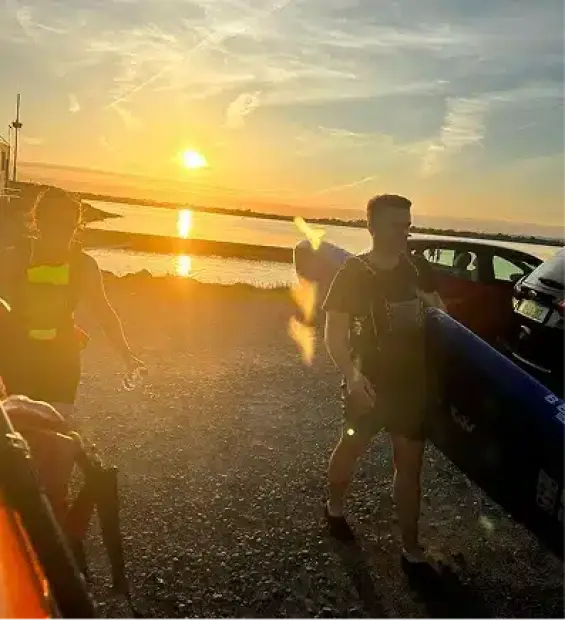 Two people carrying kayaks along a rocky shore at sunset, with the sun low in the sky reflecting on the water and cars parked nearby.